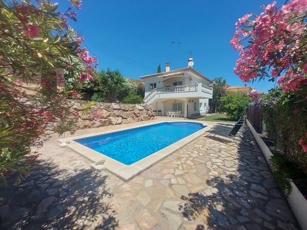 A two-story house with a swimming pool, stone patio, and surrounding greenery.