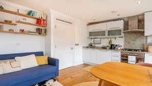 A compact kitchen with white cabinets, a stove, and a coffee maker. There's a blue sofa with a cowhide rug and a wooden dining table. - Warm and Welcoming 1BD Flat in East London (Greater London)