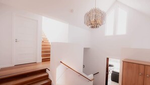 A modern staircase with wooden steps and a white handrail, leading to a room with a chandelier and a view of a balcony.