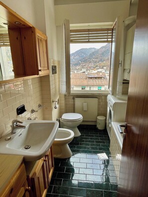 A bathroom with a sink, two toilets, a window with a view of a town and mountains, and a tiled floor. - Cozy flat Pieve di Cadore (Pieve di Cadore)