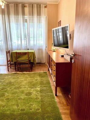 A room with a dining table, chairs, a TV on a wooden cabinet, and a green rug.