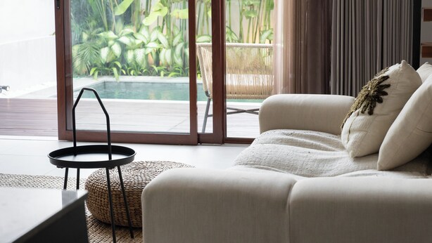 A modern living room with a sofa, a small round table, and a view of a swimming pool through large glass doors. - Lulu Villa - 2-bedrooms villa in Canggu (Canggu)