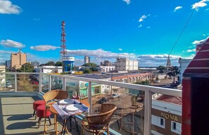 A balcony with wicker chairs and a table, overlooking a cityscape with buildings and a tall communication tower.