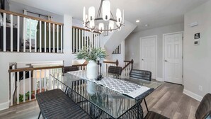 Een eethoek met een glazen tafel, zwarte metalen stoelen, een tafelkleed met een patroon, een kroonluchter en een trap met houten leuningen.