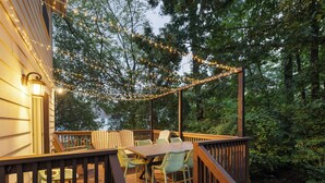 Een houten terras met een tafel en stoelen, omringd door bomen en lichtslingers.