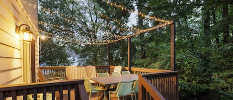 A wooden deck with a table and chairs, surrounded by trees and string lights.