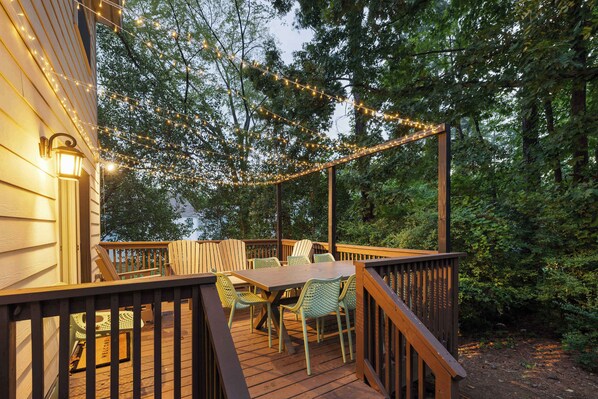 A wooden deck with a table and chairs, surrounded by trees and string lights.