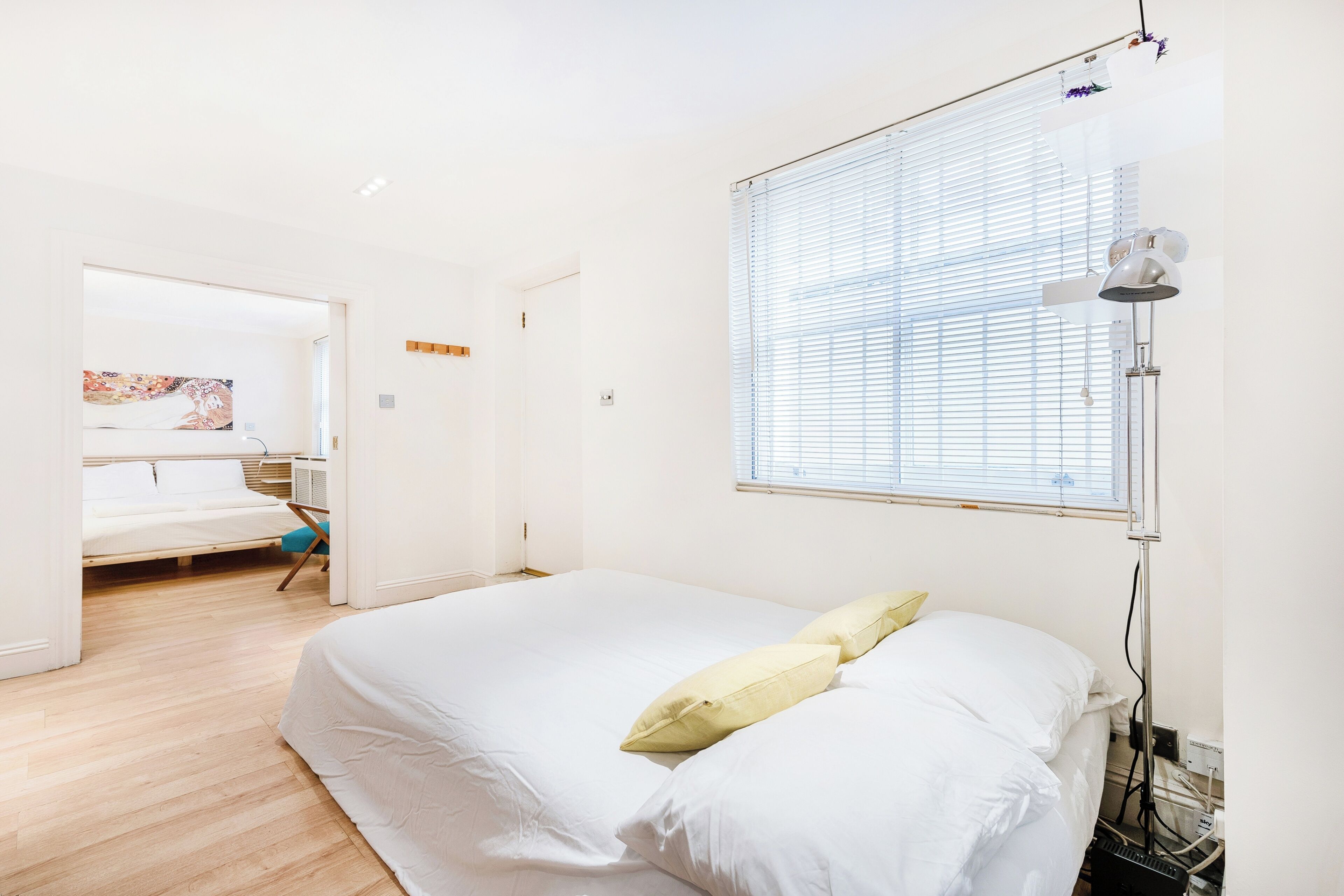 A bedroom with a bed, a bedside table, a window with blinds, and a wall-mounted lamp.