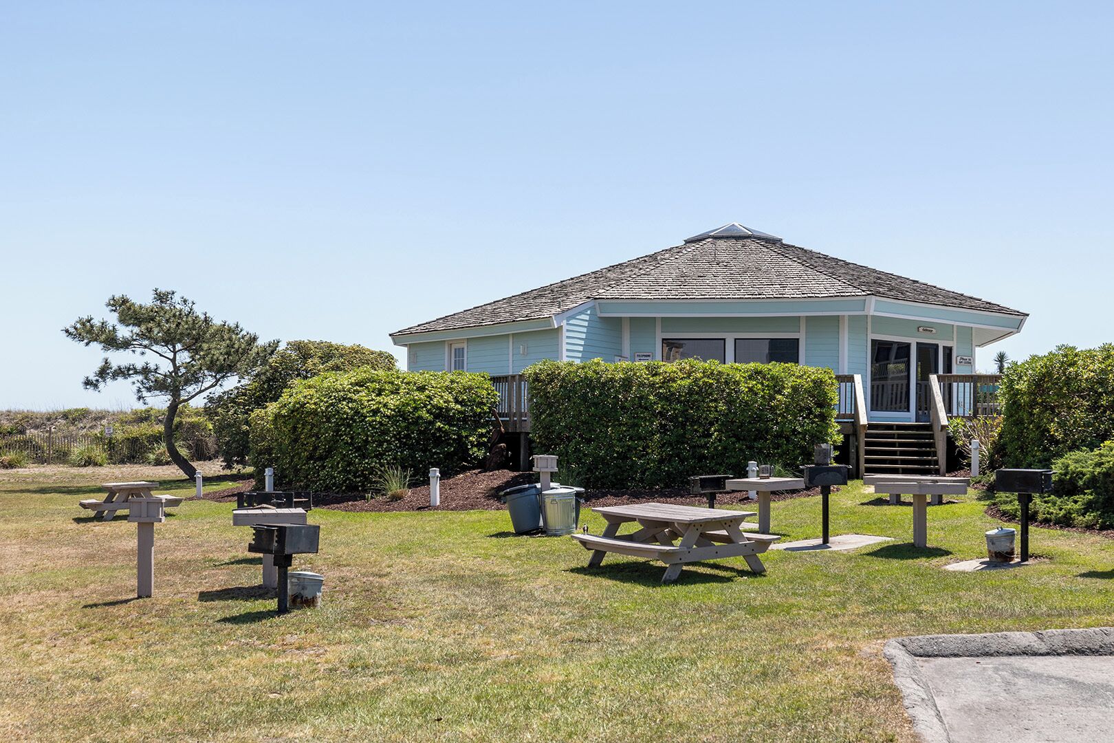 A light blue building with a covered patio area, surrounded by neatly trimmed bushes and picnic tables.