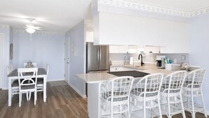 A modern kitchen with white cabinetry, a stainless steel refrigerator, and a bar area with white chairs.
