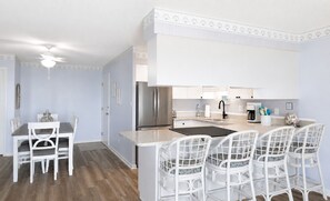 A modern kitchen with white cabinetry, a stainless steel refrigerator, and a bar area with white chairs. - Pebble Beach C-303, Almost Oceanfront 3BR 2BA (Emerald Isle)