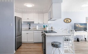 Cozinha moderna com armários brancos, geladeira de aço inoxidável, lava-louças e um espaço bar com cadeiras brancas.