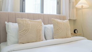 Una cama bien hecha con sábanas blancas y dos almohadas beige con textura, una mesita de noche con lámpara, y una ventana con cortinas.