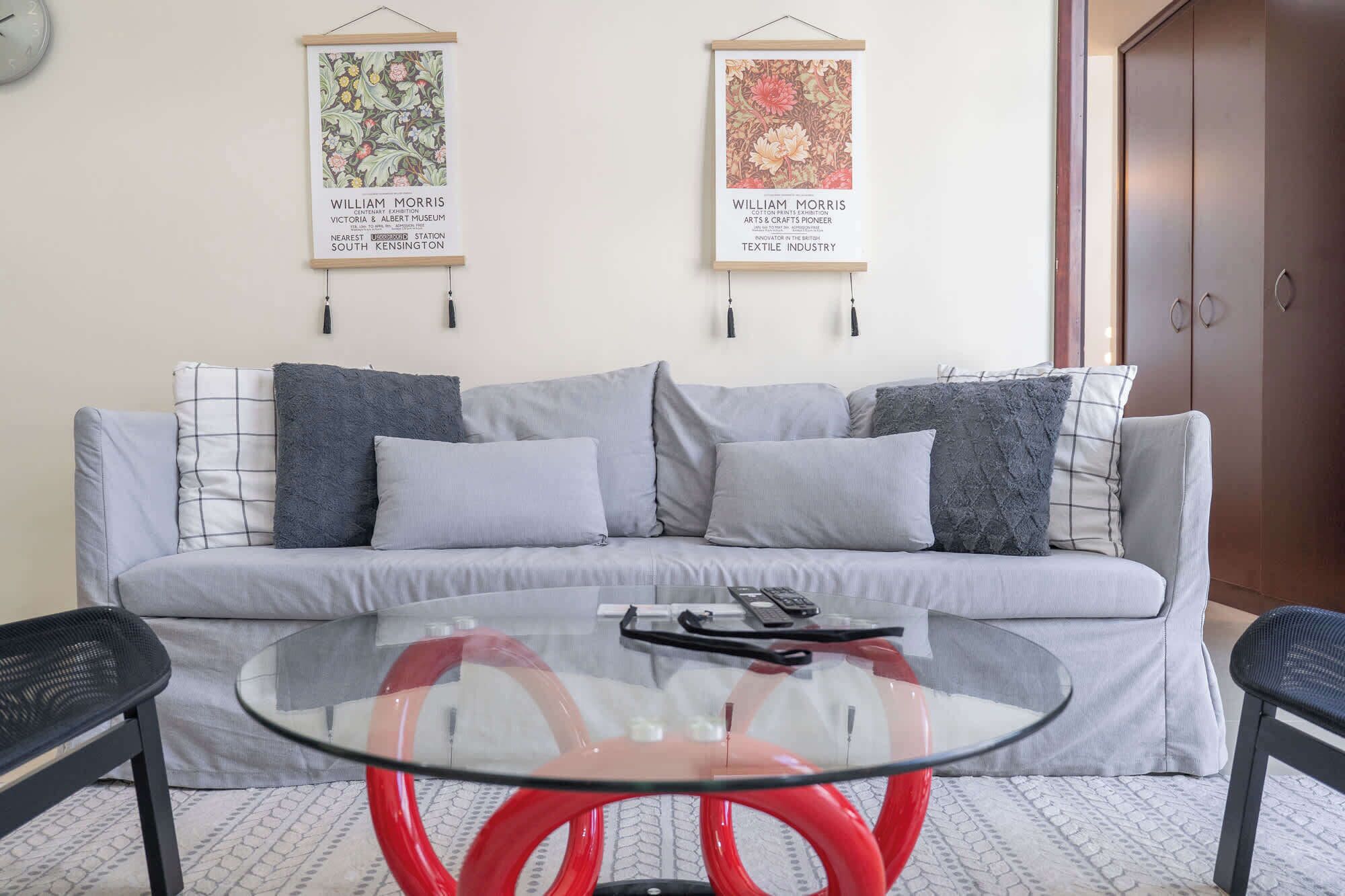 Een moderne woonkamer met een grijze bank, twee kussens, een glazen salontafel met rode poten, en twee ingelijste posters aan de muur.