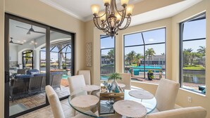 A spacious dining area with a glass table, comfortable chairs, and a decorative chandelier. Large windows provide views of the outdoor pool and palm trees. - • Sleeps 6 | 3 bedrooms |3 baths - Stars Vacation Rentals (Cape Coral)