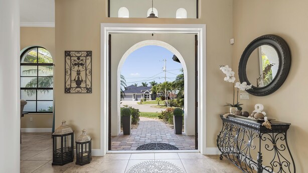 An entrance with a tiled floor, a decorative rug, a wrought iron console table, a round mirror, and a view of a garden and house through an arched doorway. - • Sleeps 6 | 3 bedrooms |3 baths - Stars Vacation Rentals (Cape Coral)