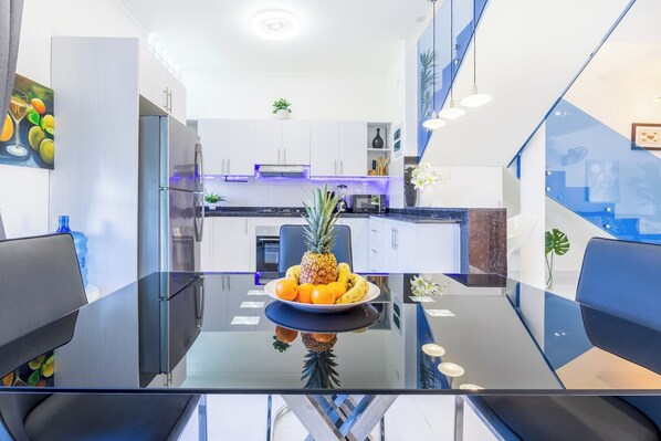 Une cuisine moderne avec une table à manger en verre, un panier de fruits et un escalier.