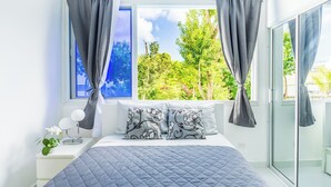Een slaapkamer met een bed, een nachtkastje, een raam met gordijnen en uitzicht op groen.