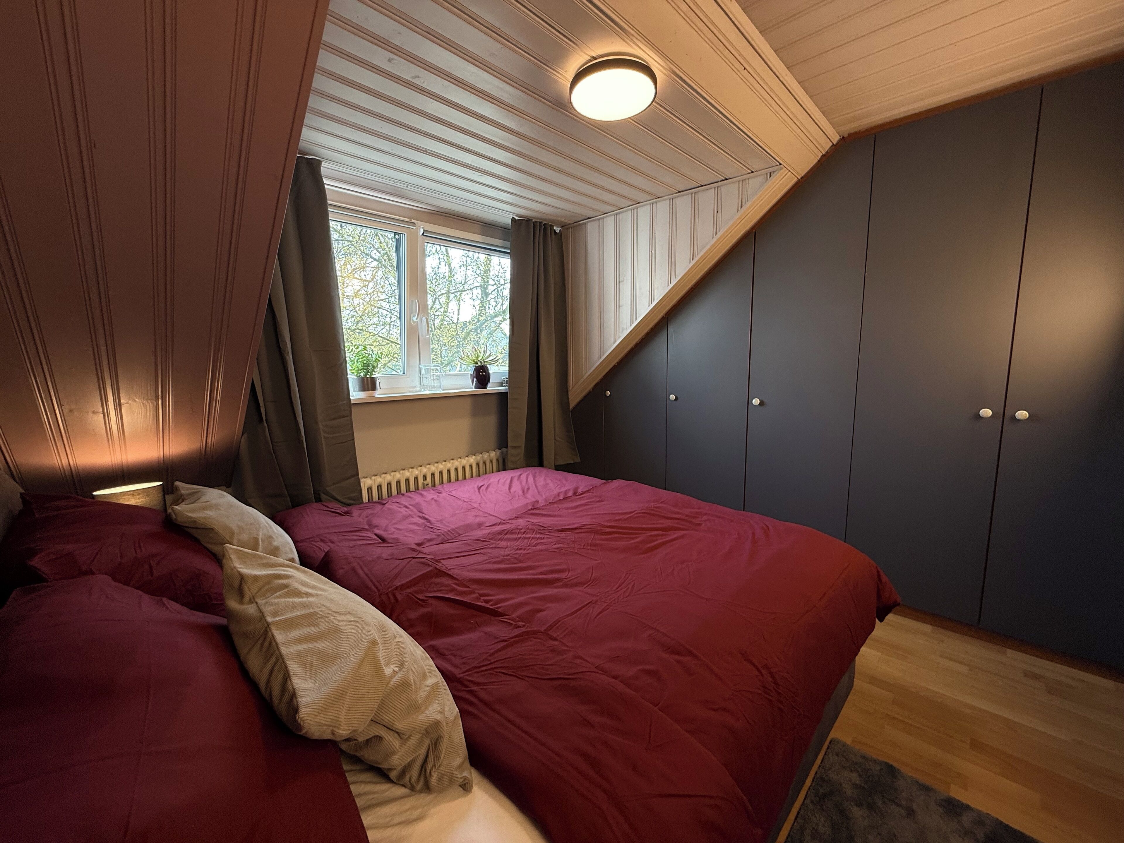 A bedroom with a bed, a window, a radiator, and dark-colored wardrobes.
