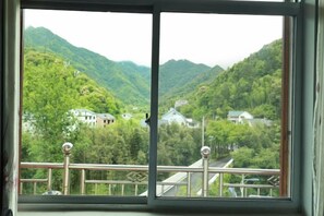 A window with a view of a mountainous landscape, a balcony with a railing, and a building in the distance. - Sanqingshan Beishan Resort Villa (Dexing)