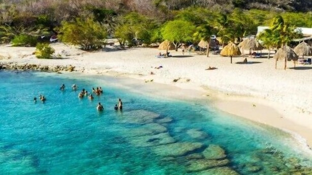 Una playa con agua turquesa cristalina, gente nadando y cabañas con techo de paja.