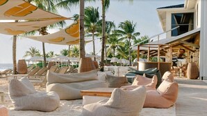 A beachside lounge area with cushioned seating and umbrellas, palm trees, and a view of the ocean.