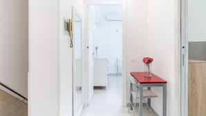 Un couloir étroit, avec une porte blanche au fond, une petite table sur laquelle est posé un vase de fleurs et une armoire blanche.