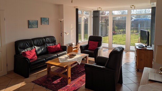 A living room with a black leather sofa, a wooden coffee table, and a red rug. There are two black armchairs, a floor lamp, and a television on a wooden stand.