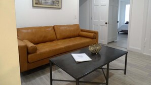 A living room with a brown leather sofa, a black coffee table with a decorative object, and a framed landscape painting on the wall. - Comfortable 3 Bed Apt in Quiet Neighborhood (Wayland)