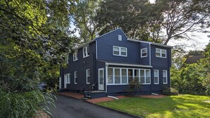 A two-story house with a dark exterior, a front porch, and a driveway leading to the entrance. - Comfortable 3 Bed Apt in Quiet Neighborhood (Wayland)