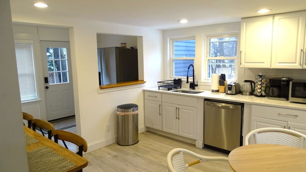 A modern kitchen with white cabinets, a stainless steel sink, and a dishwasher. There is a dining area with a wooden table and wicker chairs. - Comfortable 3 Bed Apt in Quiet Neighborhood (Wayland)