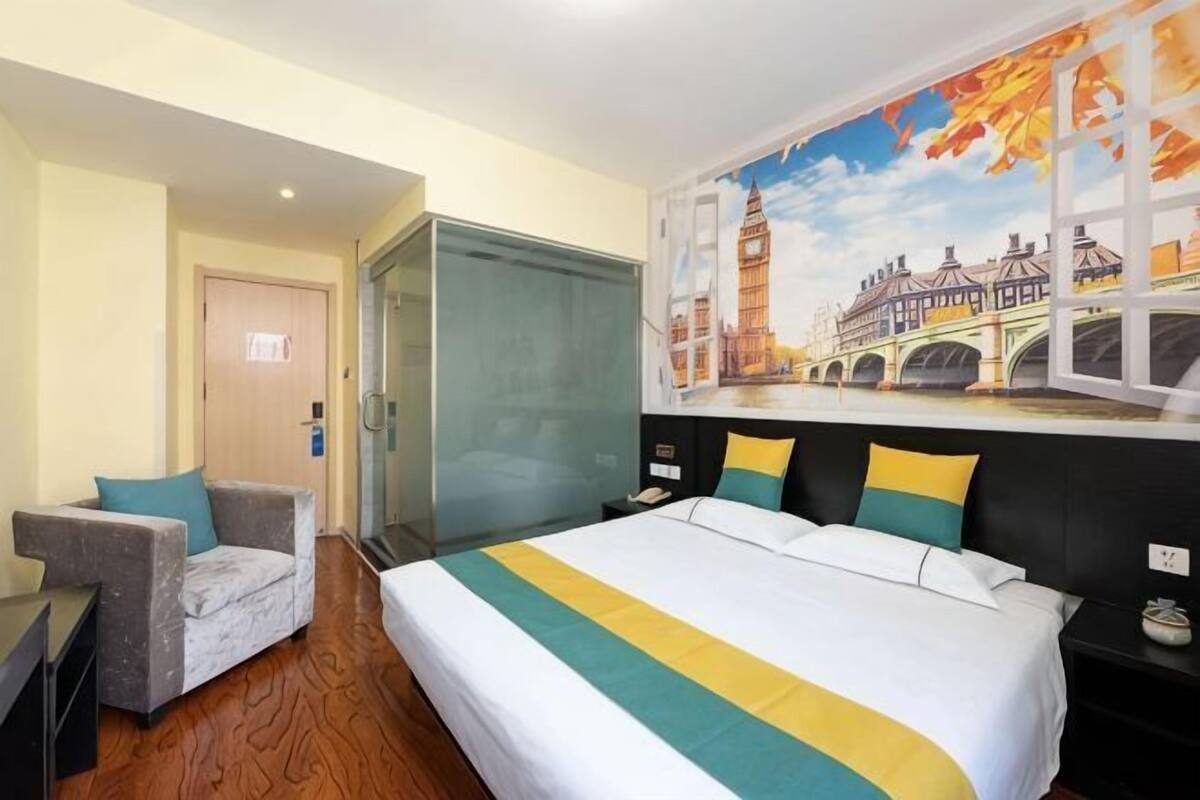 A hotel room with a bed, a chair, a desk, a glass door, and a mural of a bridge.
