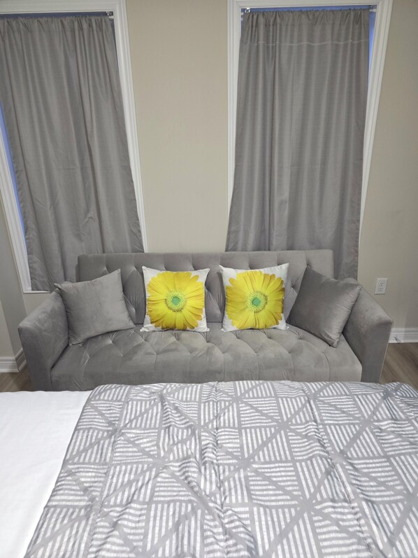 A grey tufted sofa with two yellow flower-patterned pillows, a grey and white striped throw blanket, and grey curtains.