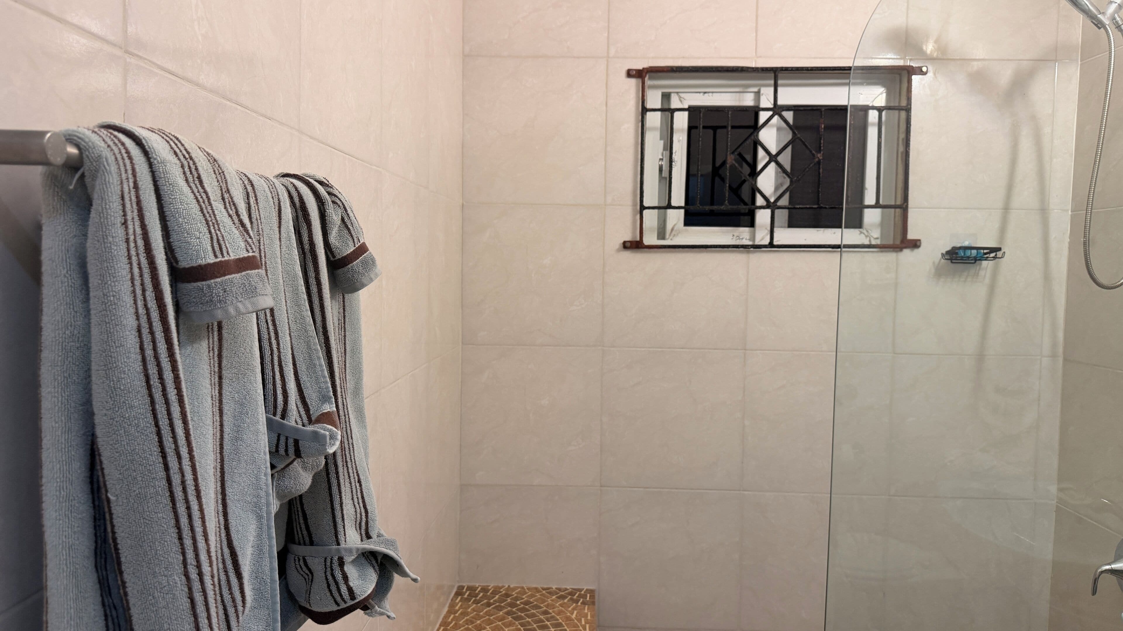A shower with a glass door, a towel rack with striped towels, and a small window with metal bars.