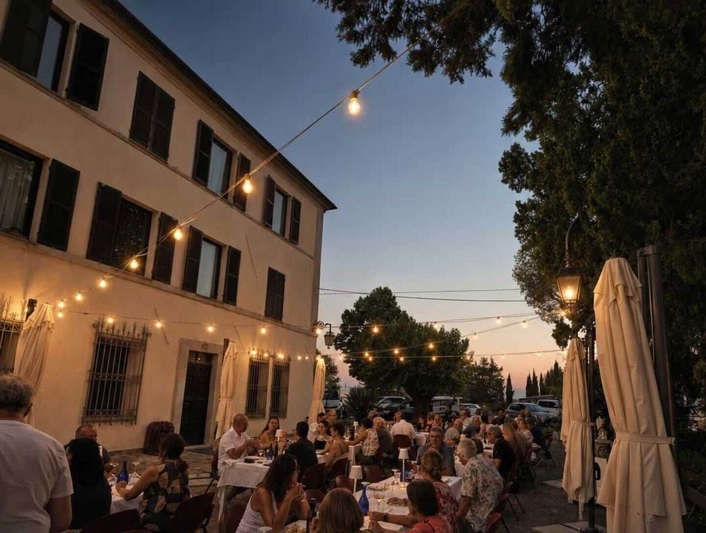 Un espacio al aire libre para comer con mesas y sillas, luces colgantes y un edificio con varias ventanas.
