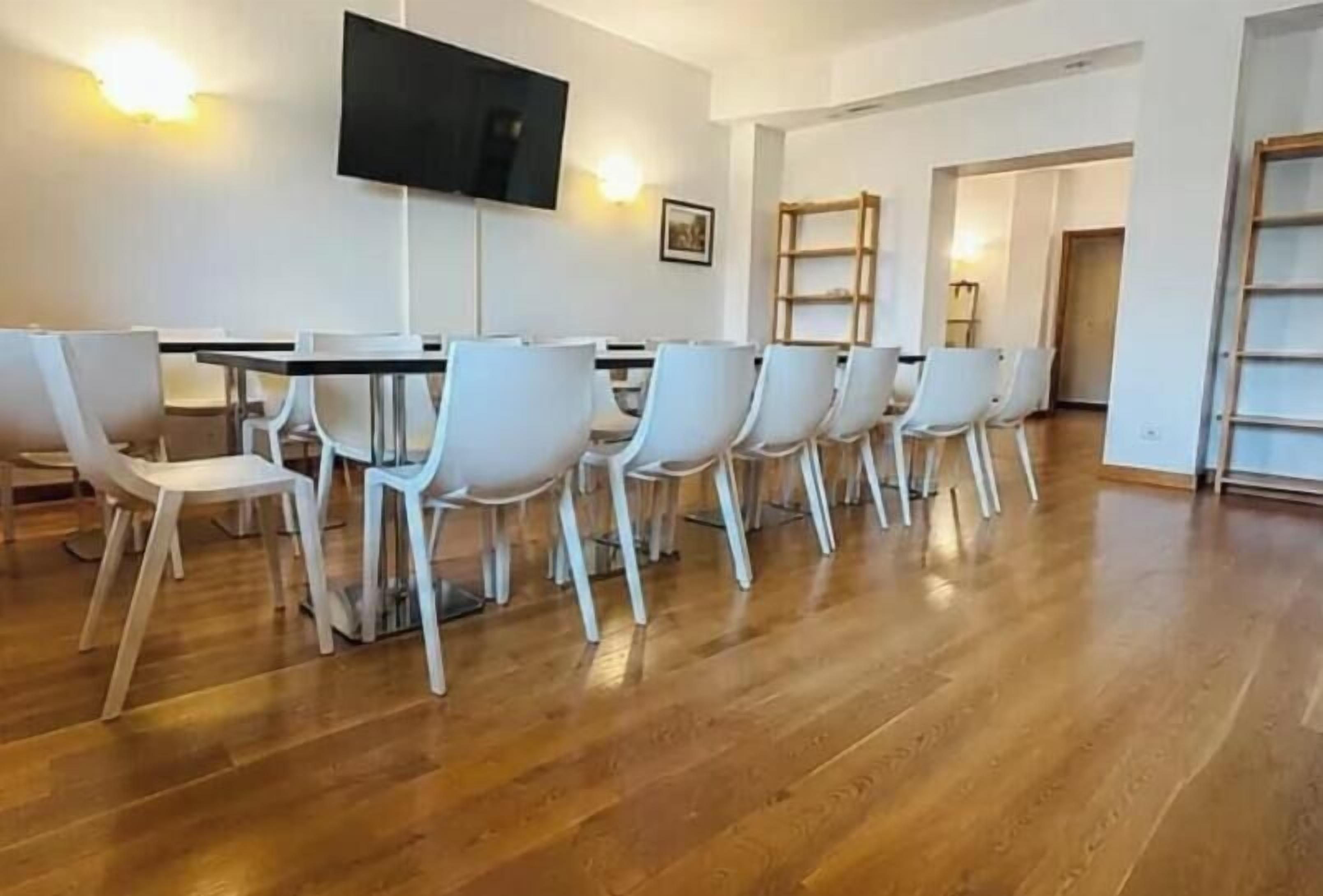 A conference room with a long table and white chairs, a flat-screen TV mounted on the wall, and a wooden floor.
