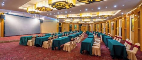 Un salón de conferencias con filas de mesas y sillas, un escenario y piso alfombrado.