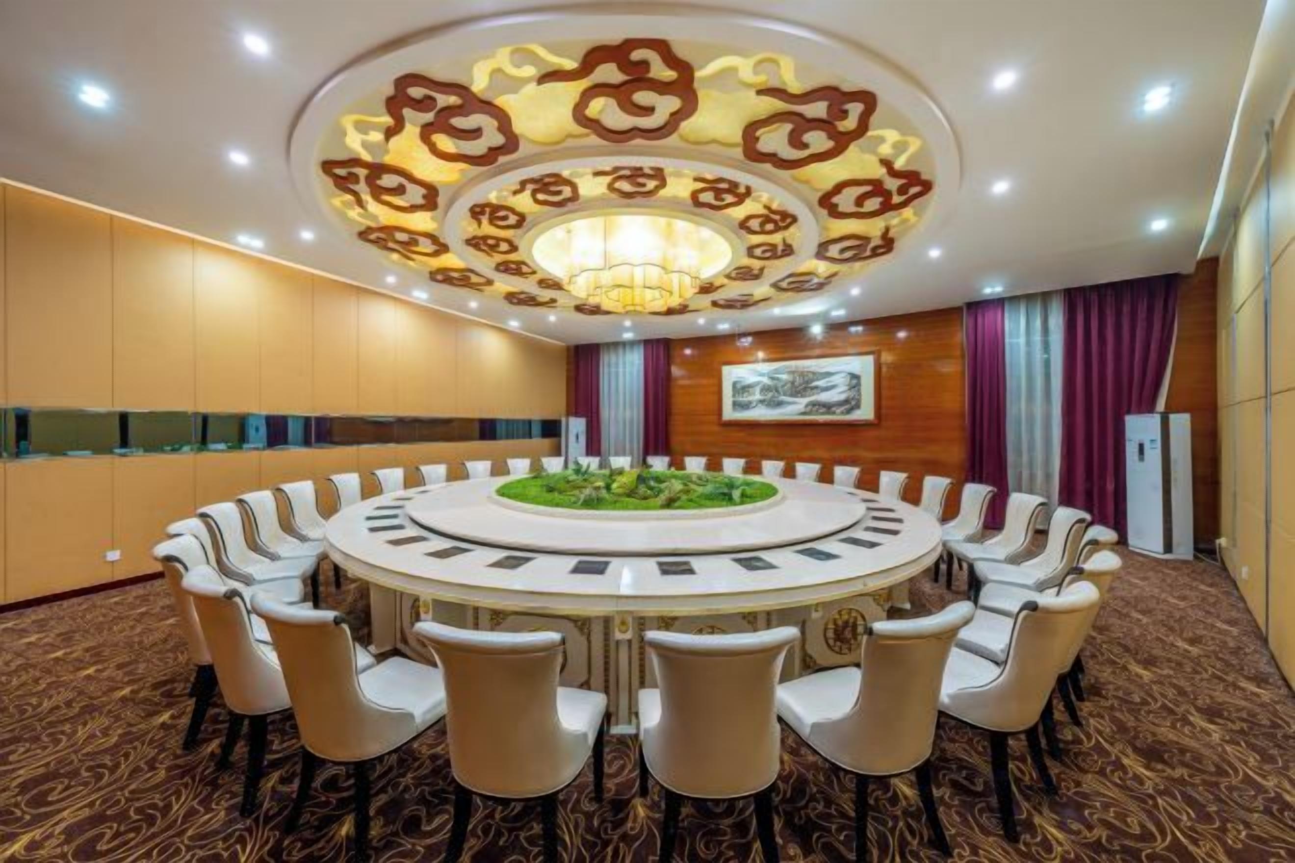 Un salón de conferencias con una mesa redonda rodeada de sillas, una araña y una alfombra estampada.