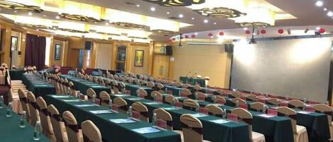 A conference room with rows of tables and chairs, a stage, and a large screen.