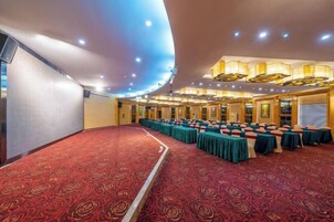 Un salón de conferencias con alfombra roja, filas de mesas con manteles verdes y un techo alto con iluminación empotrada.
