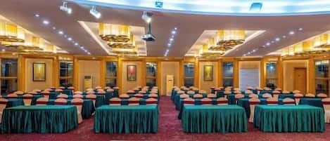 A conference room with rows of tables and chairs, a red carpeted floor, and a high ceiling with recessed lighting.