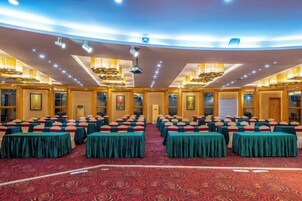 Un salón de conferencias con mesas y sillas dispuestas en filas, piso cubierto de alfombra roja y techo alto con iluminación empotrada.