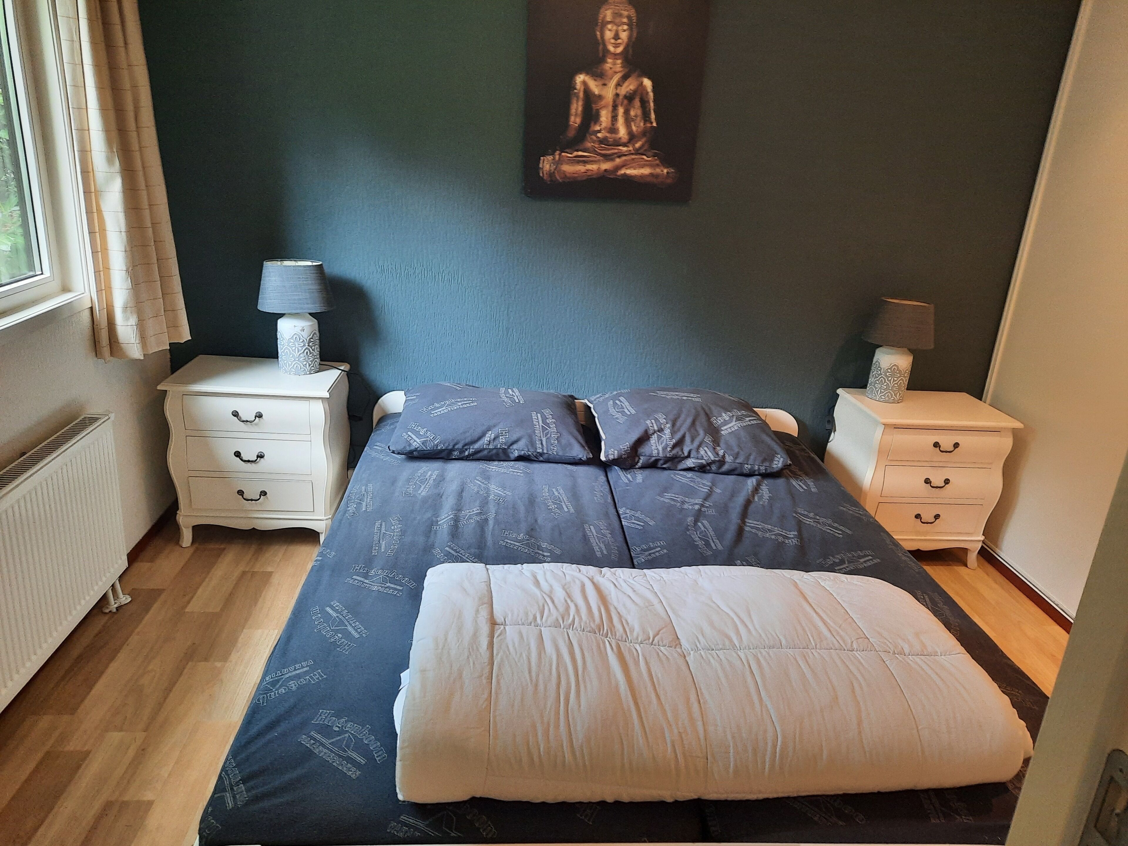A bedroom with a bed, two nightstands, a wall-mounted lamp, a window with curtains, and a Buddha statue.