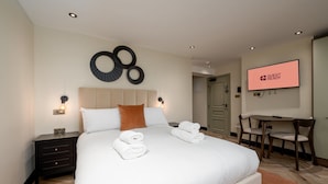 Ein Hotelzimmer mit einem großen Bett, einem Schreibtisch, einem Sessel und einem Fernseher, der die Meldung 'GUEST READY' anzeigt.