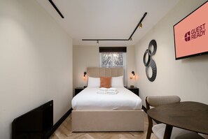 A modern hotel room with a bed, bedside tables, a window, a wall-mounted TV, and a dining area. - GuestReady - Serene getaway in Euston (London)