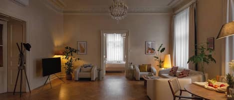 Un salón amplio con candelabro, un conjunto de sofás, una mesa de comedor con fruta y un ventanal grande.