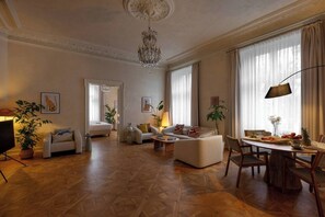 A spacious living room with a chandelier, a dining area with a table set for two, and a separate bedroom with a bed and a large window.