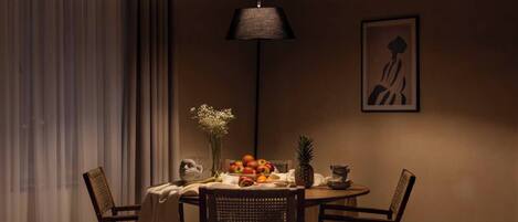 En spiseplads med et rundt bord, stole, en vase med blomster, en skål med frugt og en vægmonteret lampe.
