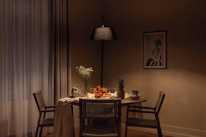 A dining area with a round table, chairs, a vase with flowers, a fruit bowl, and a wall-mounted lamp.