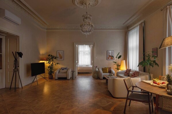 A spacious living room with a chandelier, a sofa set, a dining table with fruit, and a large window.